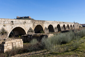 Fototapeta premium Ancient historic Roman arched stone bridge over the Guadiana river with medieval castle on a hill in the background in Medellin Spain ESP