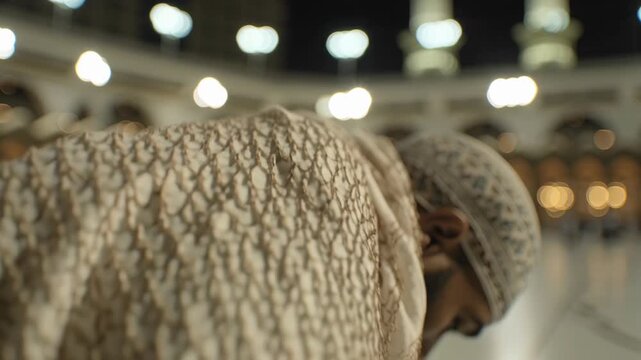 Close-up of a Muslim man in traditional attire praying at the holy mosque during night time, with blurred lights in the background for religious and spiritual
