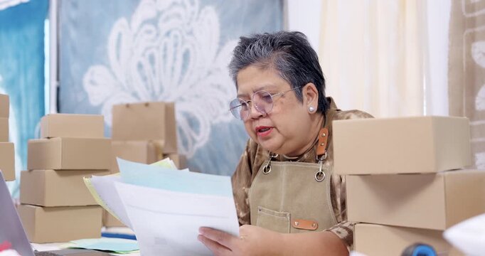 Senior woman entrepreneur leans forward in slow motion, reads paperwork, checks online order list on laptop, pauses for invoice review, plans parcel packing workflow inside home studio. home studio