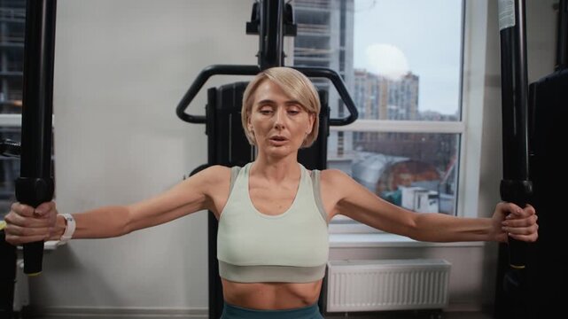 Medium shot of adult Caucasian woman in activewear performing exercise in pec deck machine in modern gym