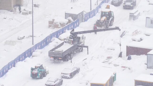 Truck mounted crane working on a construction site during heavy snowfall, winter construction work, loading materials, industrial machinery and cold weather conditions concept