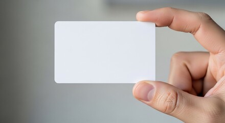 Obraz premium Hand holding a blank white business card against a blurred gray background, close-up view