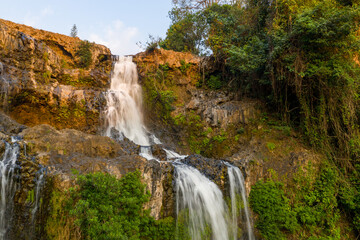 Obraz premium Warm evening light highlights the cascading waters of Tad Yuang waterfall as they flow over rugged basalt cliffs, surrounded by dense green foliage on the Bolaven Plateau in southern Laos. The