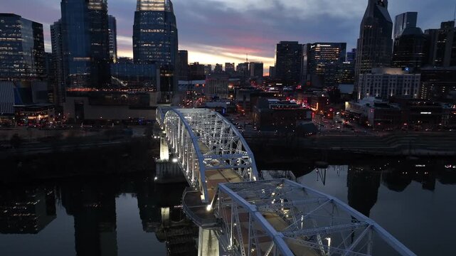 Aerial view of downtown Nashville at sunset, with illuminated bridge over cumberland river, city skyline at dusk, glowing lights, reflections on water and dramatic evening sky. Wide shot