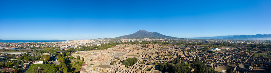 Fototapeta premium Wide panoramic perspective of the ancient ruins of Pompeii with Mount Vesuvius dominating the horizon under a clear blue sky. The image reveals archaeological textures, green spaces, and the