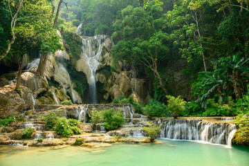 Golden sunlight illuminates the multi-level limestone cascades and turquoise pools of Kuang Si Waterfalls in northern Laos, surrounded by lush tropical forest. Vibrant green foliage and flowing water © Florent