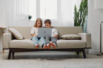 Fototapeta premium Two children sitting barefoot on a beige sofa using a laptop together in a bright living room with natural light and plants. Concept of technology, learning, and family bonding.