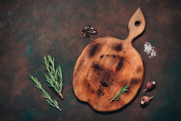 Empty cutting wooden board with spices and herbs on dark grunge background, culinary backdrop, top view, flat lay, copy space.