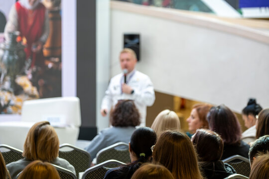 Doctor in white coat delivers talk to seated audience in busy conference area