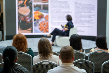 Obraz premium Audience sits facing stage during conference session watching speaker present large screen images and text.