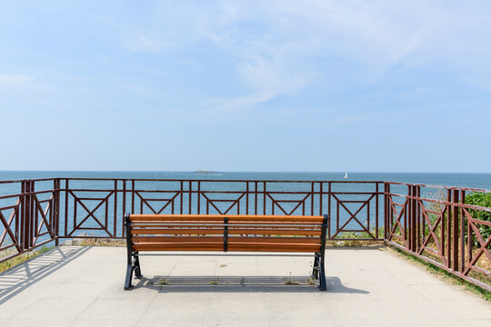 Wooden bench on a concrete terrace with metal railing facing a tranquil sea, under a clear sky with distant sailboats and an island on the horizon.
