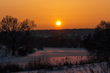 Obraz premium Majestic winter sunset warms a tranquil frozen river, reflecting golden hues on crisp snow. Bare trees silhouette against a brilliant sky, with a hint of distant dwellings