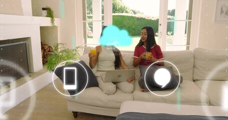 Naklejka premium Sharing two women sipping orange juice and using silver laptop on sofa in bright living room