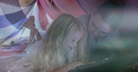 Lying two girls watching tablet in DIY blanket fort, wearing pajamas, with pillows and fairy lights