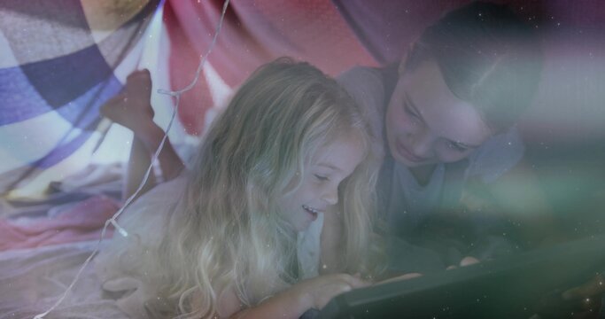 Lying two girls watching tablet in DIY blanket fort, wearing pajamas, with pillows and fairy lights