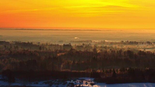 Distant aerial of snow covered city edge smokestack rising under golden sunrise sky and winter haze, forest treetops diffuse light