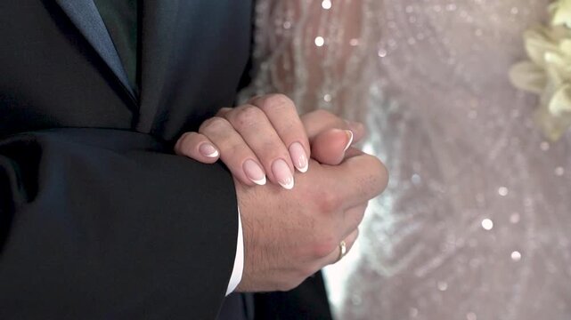 A couple stands together holding hands in a wedding ceremony. The venue has flowers around them as they exchange vows. Guests are present and celebrating their union.