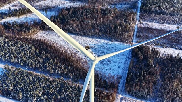 Aerial tilt down flyover above a motionless wind turbine surrounded by snowy forest clearings during beautiful winter sunrise