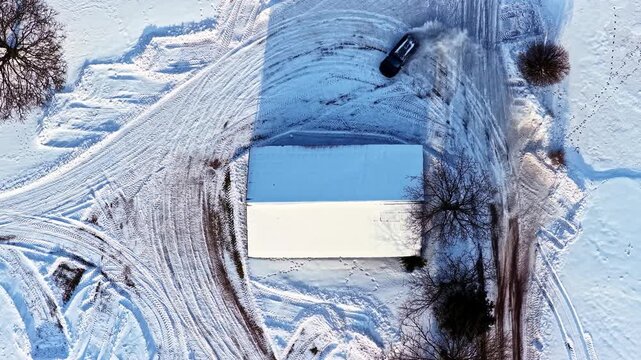 Aerial top down view of a car drifting in circles kicking up snow around a large snow-covered house at beautiful winter sunrise