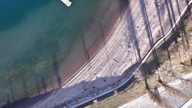 Minimalist bird's-eye view of Lake Walensee's coast. Turquoise water meets a pebbled beach with long, dramatic winter shadows from lakeside trees near Walenstadt.