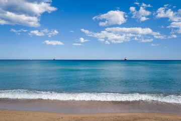 Peaceful view of a sandy shoreline with small white waves meeting clear blue water under a sky dotted with fluffy clouds near Lotzorai, Sardinia. Two distant boats add interest to the tranquil