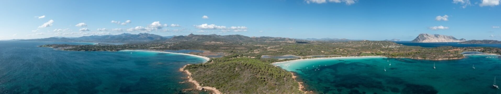 Expansive drone panorama of Capo Coda Cavallo near San Teodoro, Sardinia, showing turquoise bays, sandy beaches, lush greenery, and distant mountains beneath a clear blue sky.