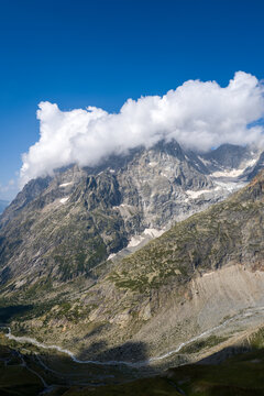 Massive rocky mountain slopes with patches of green vegetation rise sharply beneath a thick layer of white clouds, set against a deep blue sky in the Glacier de Pre de Bar and Mont Dolent area.