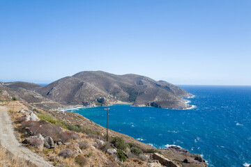 Fototapeta premium Dirt path runs along a rugged hillside overlooking a vivid blue bay and rocky mountains on the Mani Peninsula, Greece. Windswept vegetation and sparkling sea create a wild, untouched Mediterranean