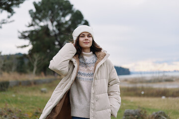 Young woman in a warm beige winter coat stands outdoors by a lakeside, wearing a knit sweater and...
