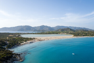 Obraz premium Sweeping aerial perspective of Porto Giunco reveals a sandy beach curving between turquoise sea and tranquil lagoon, framed by lush greenery and distant mountains under a bright sky.