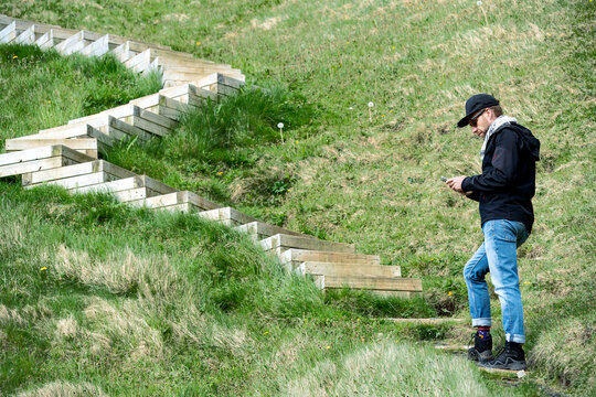 View of a person pauses on a winding staircase amidst vibrant green slopes, absorbed in a mobile device, Vestmannaeyjabaer, Vestmannaeyjabaer, Iceland.
