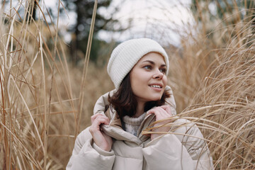 Woman wearing a warm coat and white beanie stands among tall grasses in a chilly outdoor setting,...