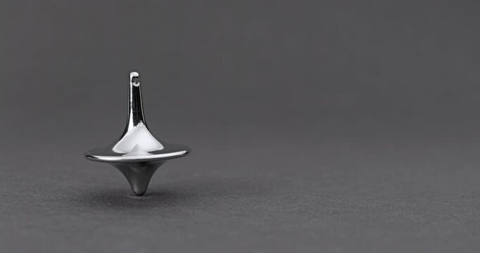 Metallic Spinning Top Falling Over. Linear footage of a shiny chrome top in steady rotation, progressively wobbling with increasing tilt until it fully topples on a matte gray surface.