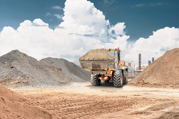Obraz premium Loader picks up sand and transports it on a construction site surrounded by piles of earth and buildings in the background. Work is ongoing during daytime