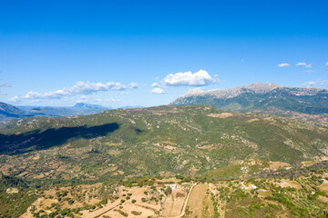 Obraz premium Wide aerial perspective of rolling green hills and rugged mountains near Orgosolo, Sardinia, with scattered clouds and sunlit farmland creating a sense of open space and tranquility.