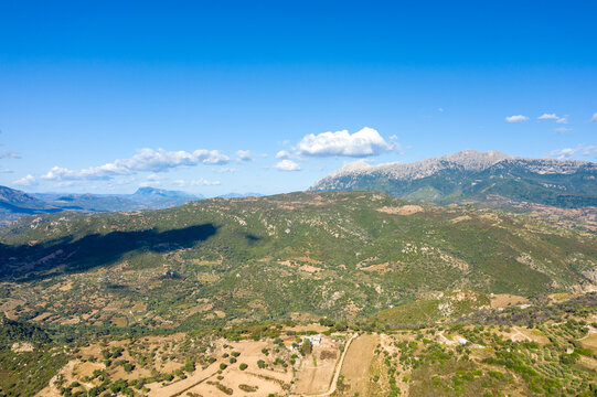 Wide aerial perspective of rolling green hills and rugged mountains near Orgosolo, Sardinia, with scattered clouds and sunlit farmland creating a sense of open space and tranquility.