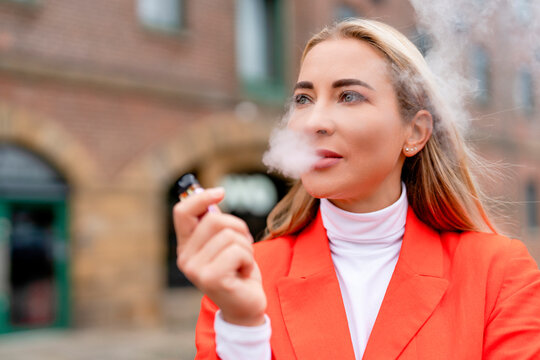 Woman smokes a vape while standing outside with brick buildings in the background during daytime