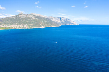 Fototapeta premium Wide aerial view of the deep blue Mediterranean Sea meeting the rugged green mountains and coastline near Lotzorai, Sardinia. The scene is bright and tranquil with a clear sky and subtle waves.