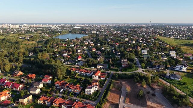 Drone pullback shot over Gilužiai village showing houses, undeveloped plots, a water body and the city skyline in the distance near Vilnius, Lithuania.