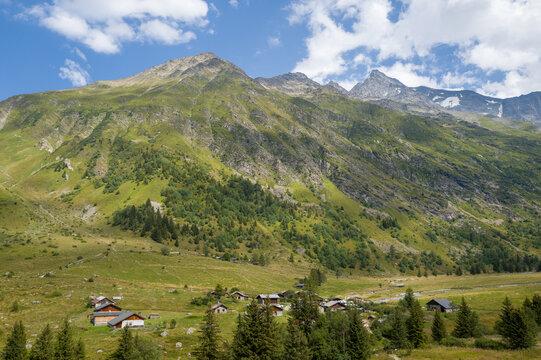 Scattered wooden chalets sit in a lush green valley at the foot of steep, rocky mountains, surrounded by evergreen trees under a partly cloudy summer sky in the French Alps.