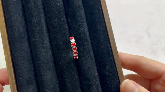 Close-up view of a hand holding a silver ring adorned with red heart shapes, displayed on a black velvet jewelry holder with multiple compartments in a bright, neutral setting