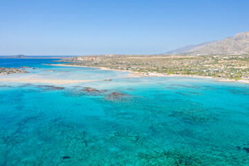 Expansive panorama of Elafonisi beach in Crete, featuring vivid turquoise water, sandy shallows, and rugged hills under a cloudless sky. The clear light reveals underwater textures and the natural