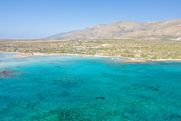 Obraz premium Expansive aerial perspective of Elafonisi beach in Crete, showcasing clear turquoise sea, sandy shoreline, and rugged hills beneath a cloudless blue sky. The vivid colors and open landscape evoke a