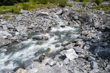 A fast-flowing alpine river carves through a rugged bed of gray stones and boulders in Cirque du Fer-a-Cheval. Bright daylight highlights the textured rocks and lively water in this dramatic mountain