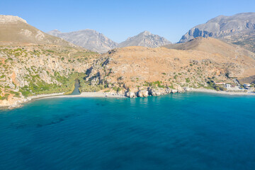 Aerial view of Preveli beach in Crete, where a winding river lined with lush greenery meets a sandy shoreline and vivid blue sea, framed by rugged mountains and sunlit rocky cliffs under a clear sky.