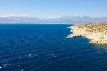 Expansive view of rugged limestone coastline meeting the deep blue Mediterranean near Matala, Crete, with distant hazy mountains under a clear sky. The scene evokes openness, wild textures, and