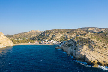 Fototapeta premium Aerial view of Matala on Crete, showing a small seaside village nestled between dramatic limestone cliffs and deep blue water, with sunlit hills in the background under a clear sky.