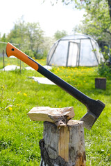 An axe stuck into a wooden stump in a bright green garden with a greenhouse in the background demonstrates gardening tools and nature. Vertical background of country life.