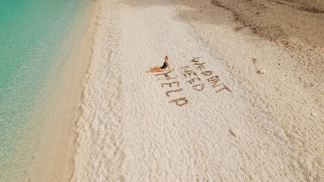 Woman relaxing on quiet Kefalonia shore beside playful phrase we dont need help in sand.