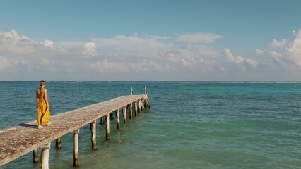 Obraz premium Woman in yellow dress walks along wooden pier over a calm tropical sea waters.
